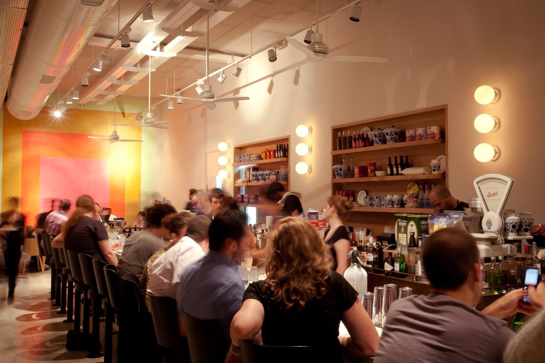 Interior of Mizlala restaurant in Tel Aviv with diners seated at a curved wooden bar, shelving with bottles, and colorful ambient lighting.