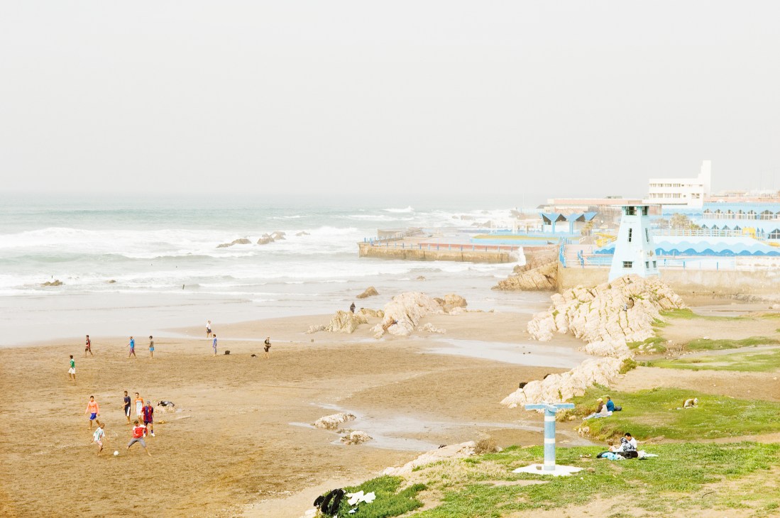 Casablanca beach with children playing, rocky outcrops, and blue beach clubs lining the shore.