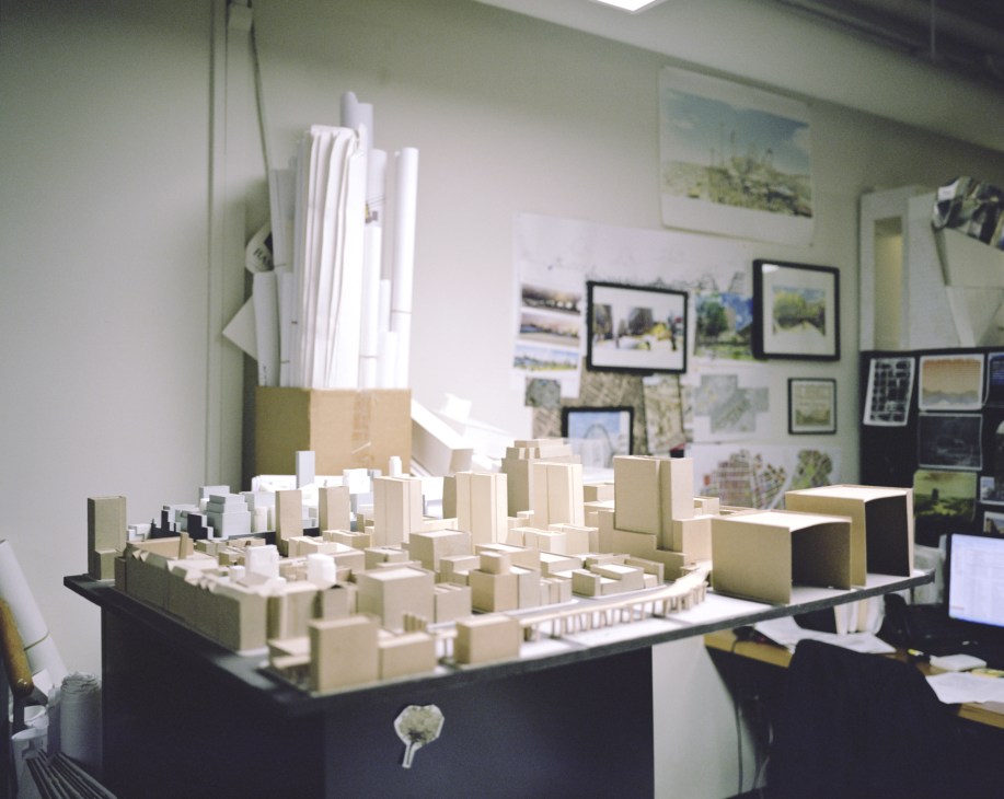 Architectural models of buildings displayed on a table in an urban design office with sketches and reference images on the walls.