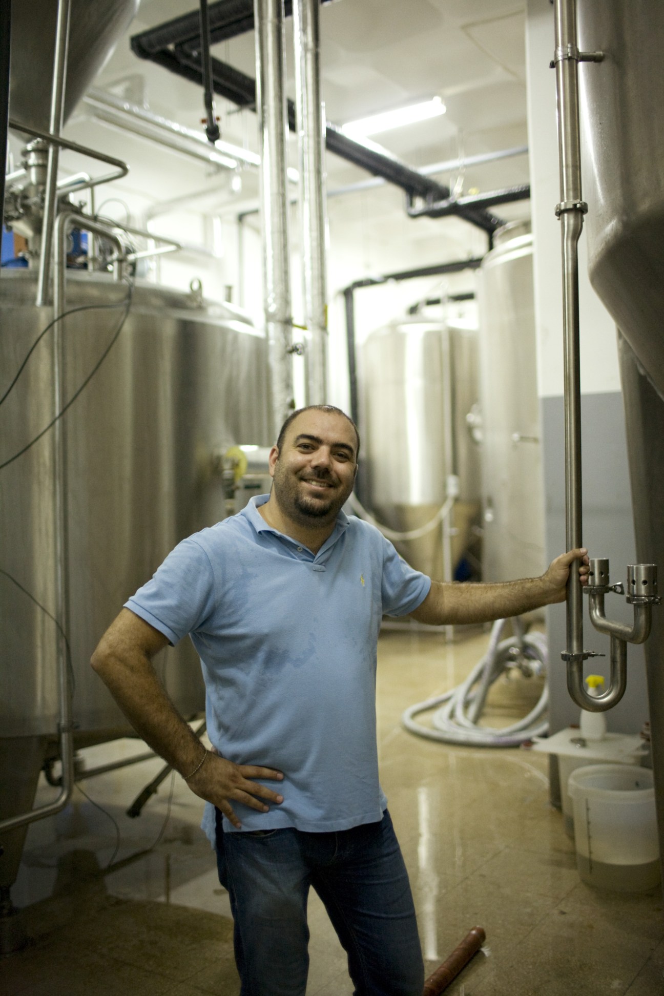 Man in blue shirt smiling in brewery surrounded by large metal fermentation tanks and pipework.