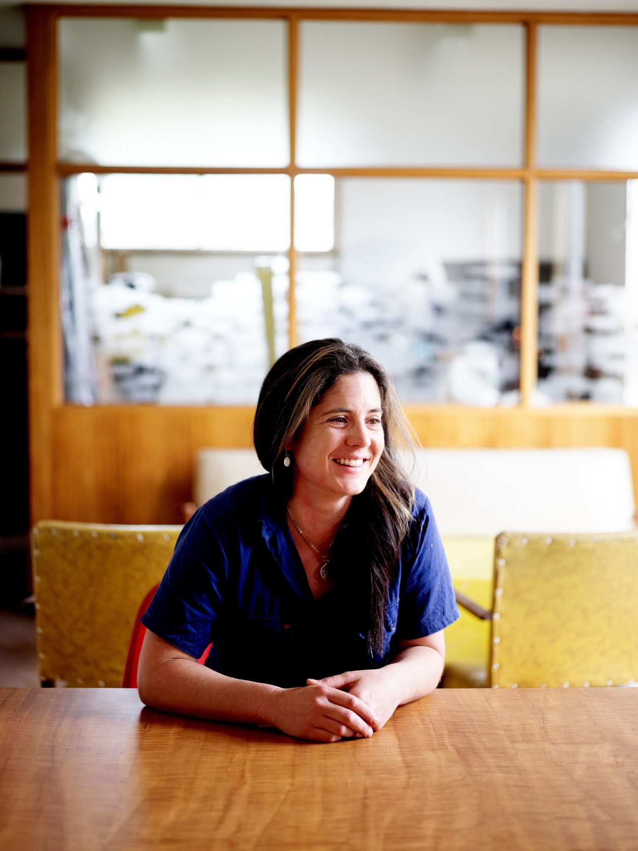 Laura McCusker sits at a wooden table in her workshop, smiling at the camera.