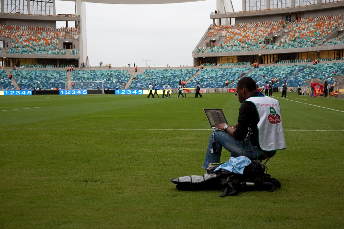 Photographer sitting on a football pitch with a laptop, filing pictures during a World Cup pre-match event at a stadium.
