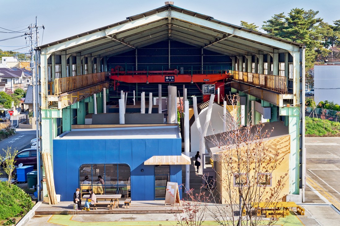 Aerial view of Newland community space in Kumagaya showing converted industrial warehouse with blue storefront, red crane machinery visible, wooden walkways, and planted greenery.