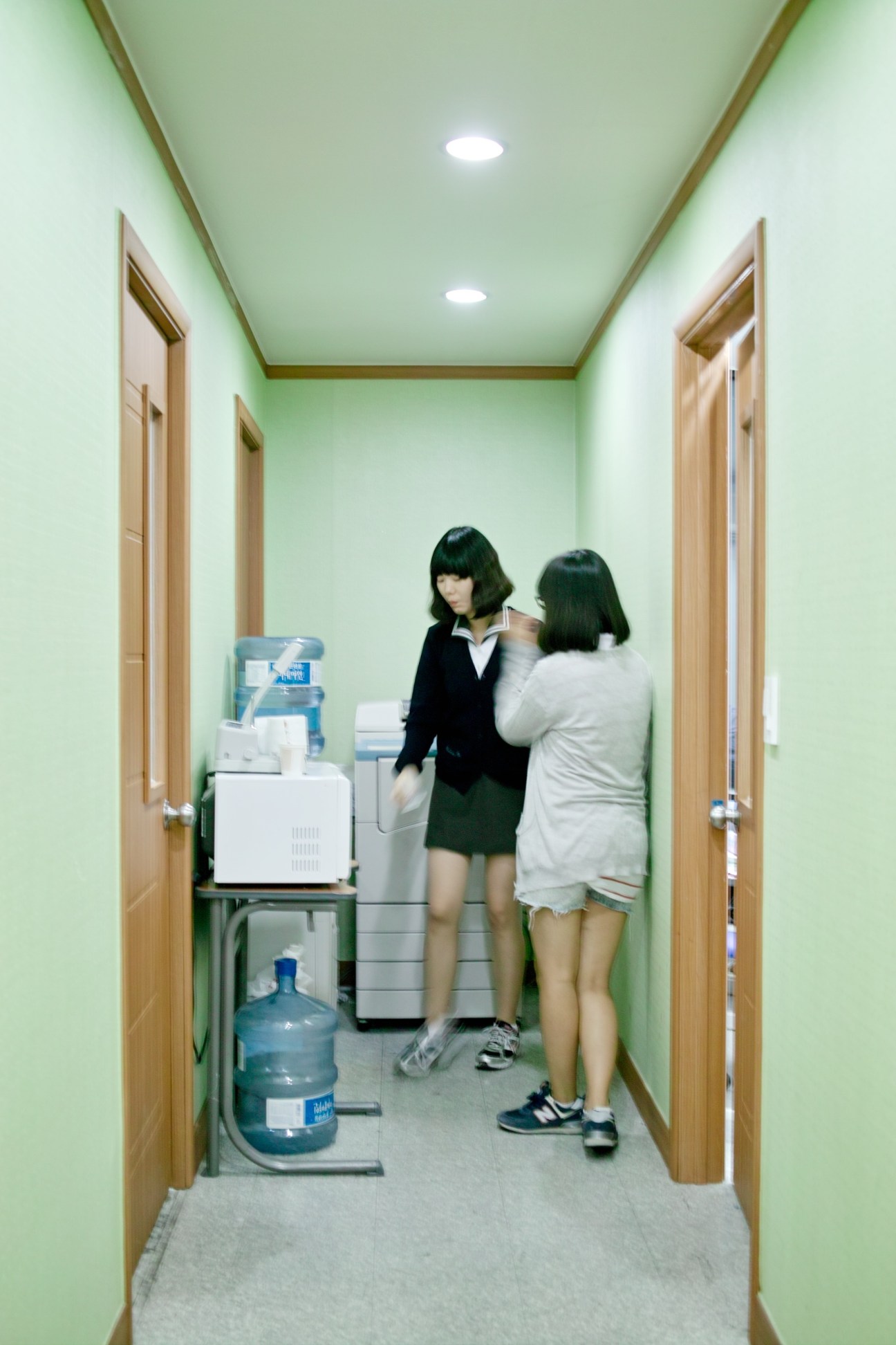 Two teenage girls in a narrow hallway with green walls and wooden doors, one wearing a school uniform.
