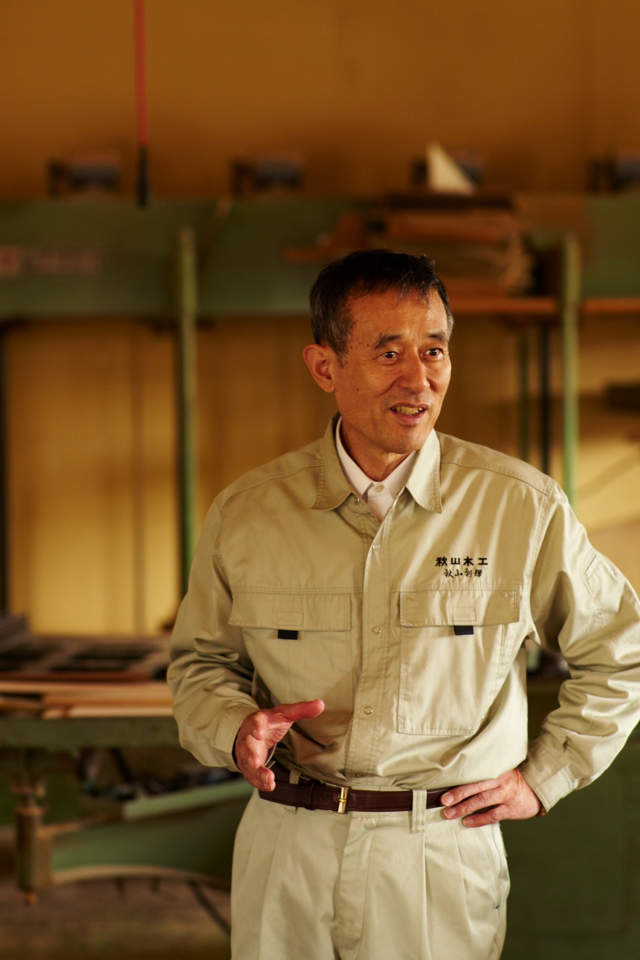Toshiteru Akiyama, founder of Akiyama Mokko furniture workshop, standing in his workshop wearing a beige work shirt with the company logo.