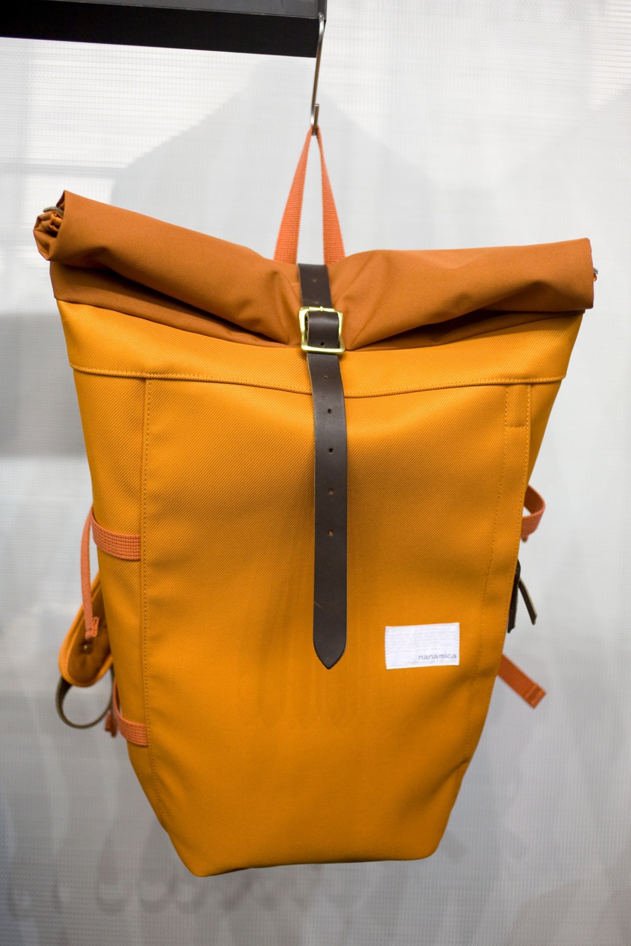 Yellow Nanamica backpack with rolled-top closure and orange straps hanging from a display hook.