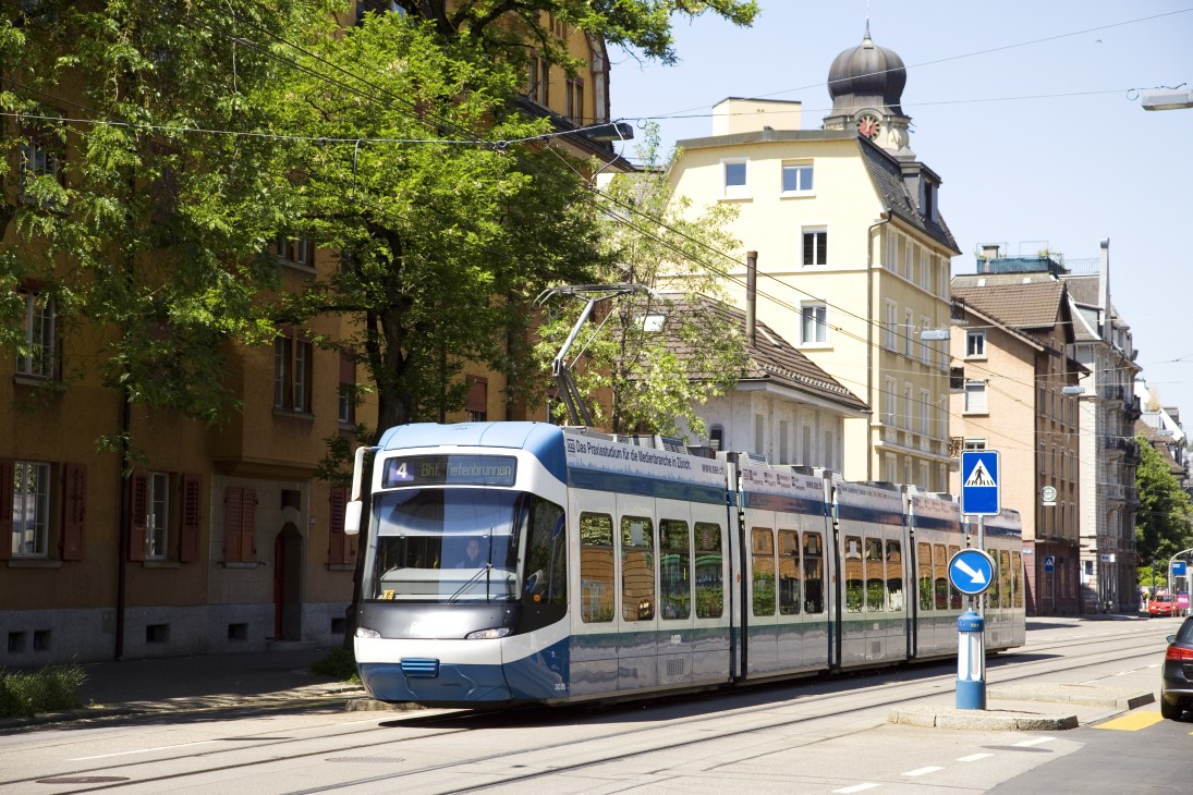 Modern tram running through a tree-lined street in Zurich with historic buildings.