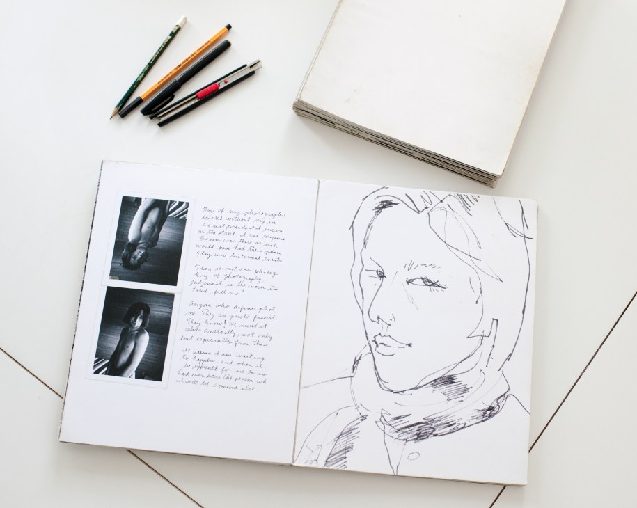 Open sketchbook with black and white photographs and pen drawings of a face, alongside colored pens and blank notebook.