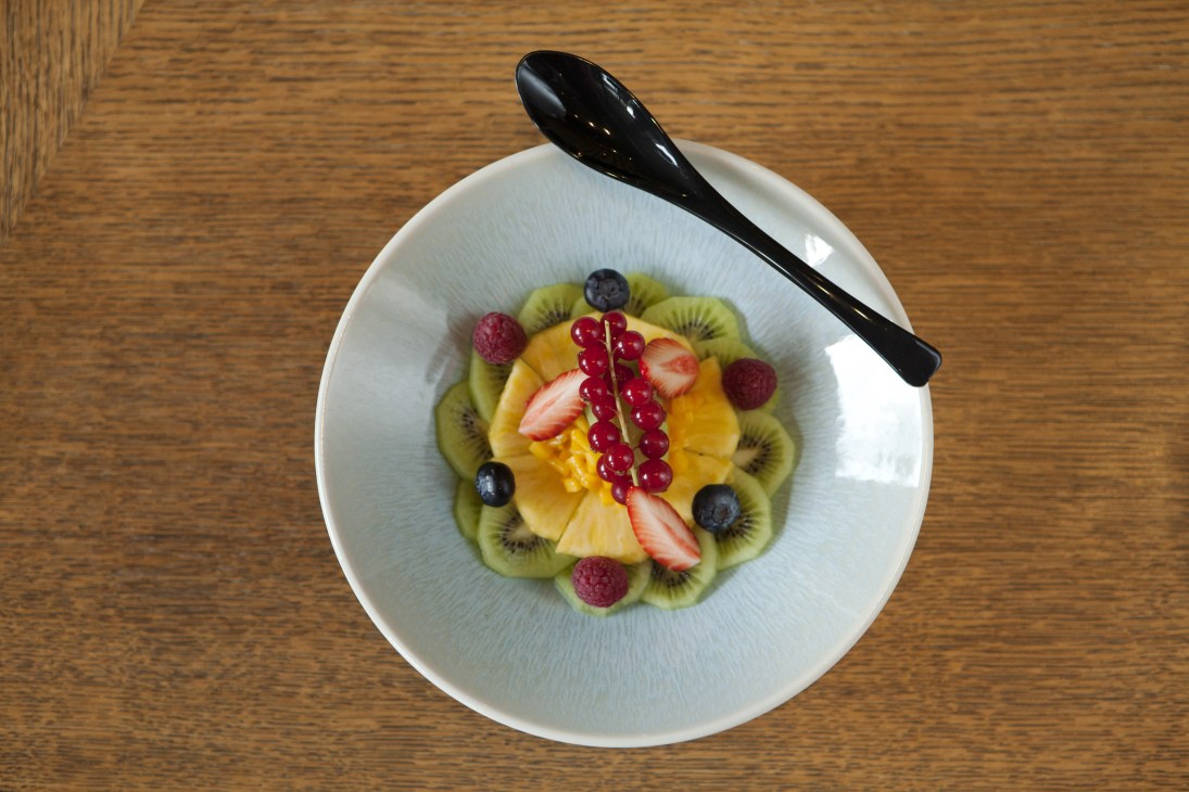 Kudamono fruit salad with kiwi, mango, pineapple, strawberry, raspberries, blueberries and redcurrants arranged on a white plate with a black spoon.