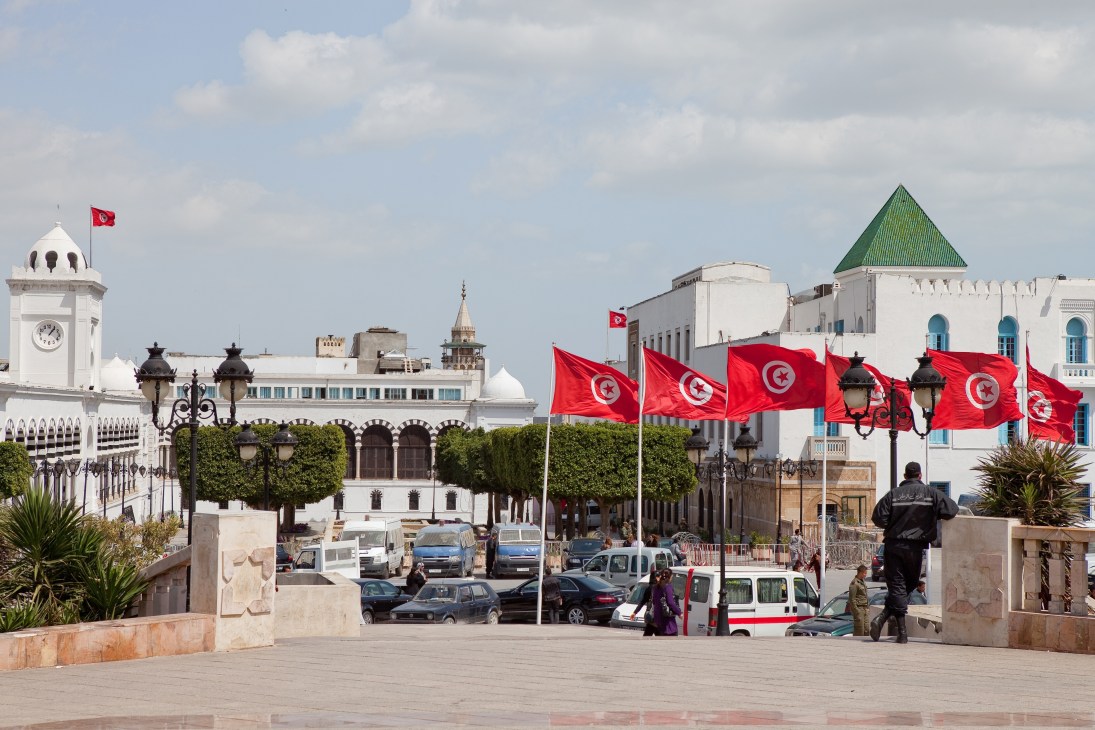 Tunisian flags displayed in Place du Gouvernement with white colonial-era buildings and a green-roofed mosque in background.