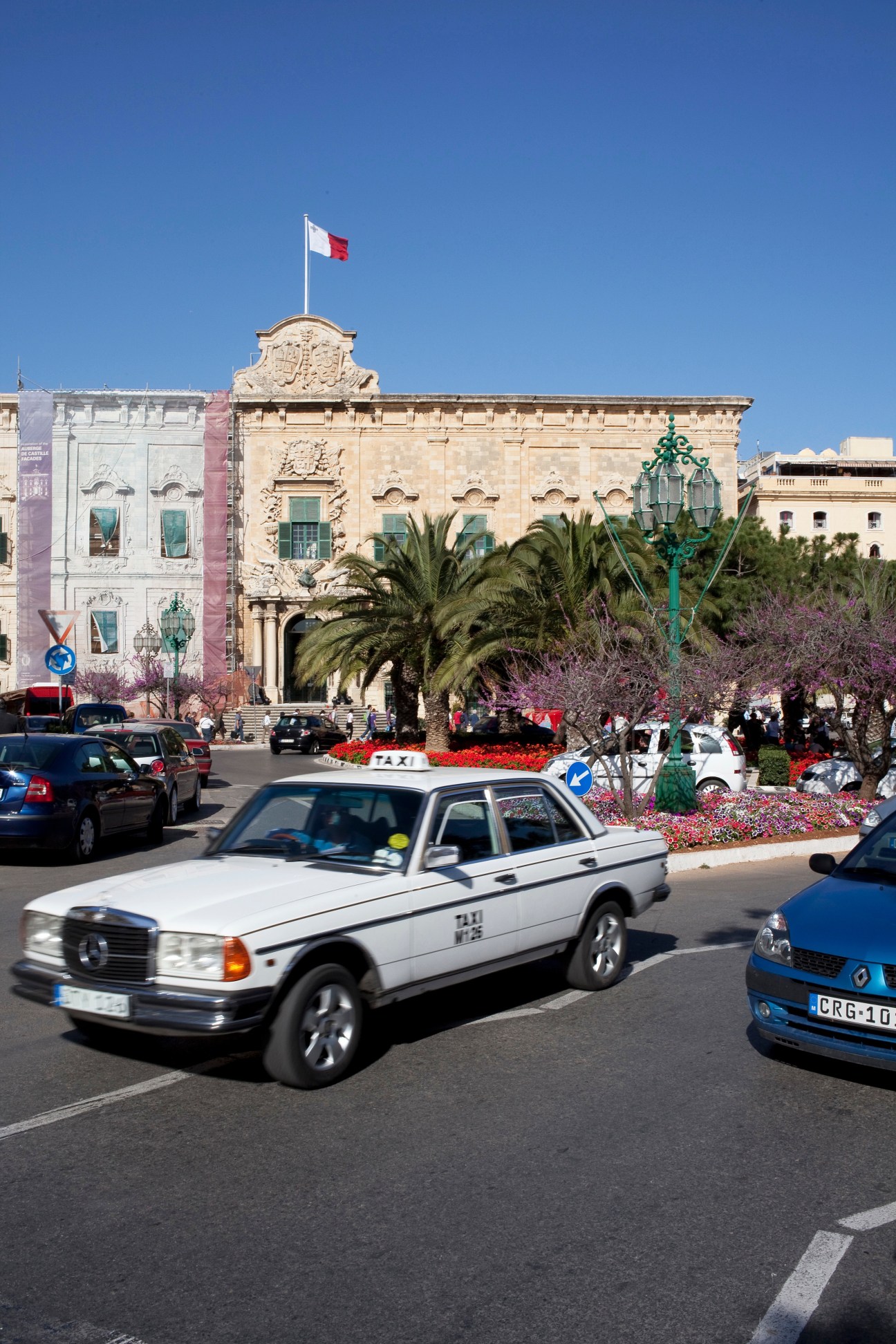 Auberge de Castille building exterior in Valletta with Maltese flag, featuring classical architecture and a taxi in the foreground.