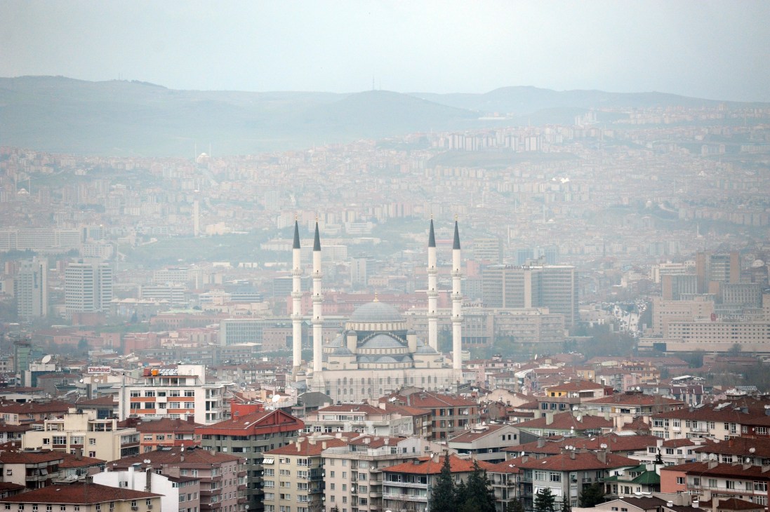 Aerial view of Ankara showing a white mosque with twin minarets surrounded by residential buildings and hills in hazy conditions.