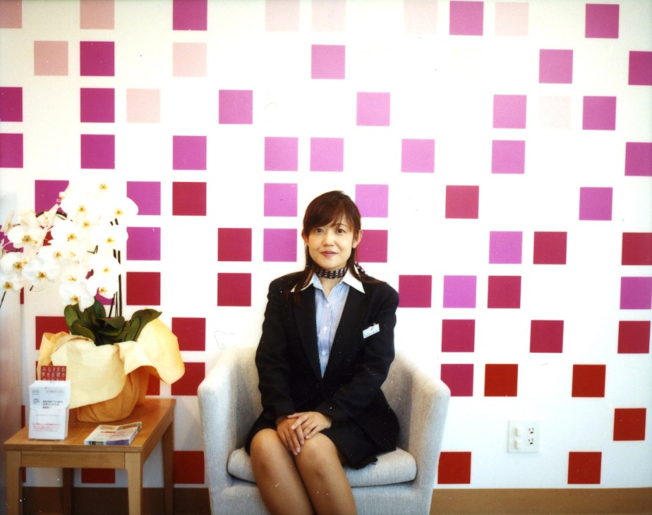 Bank employee Rie Takanashi sits in a light grey chair against a wall of colorful pink and red squares at Sugamo Shinkin Bank's Niiza branch.