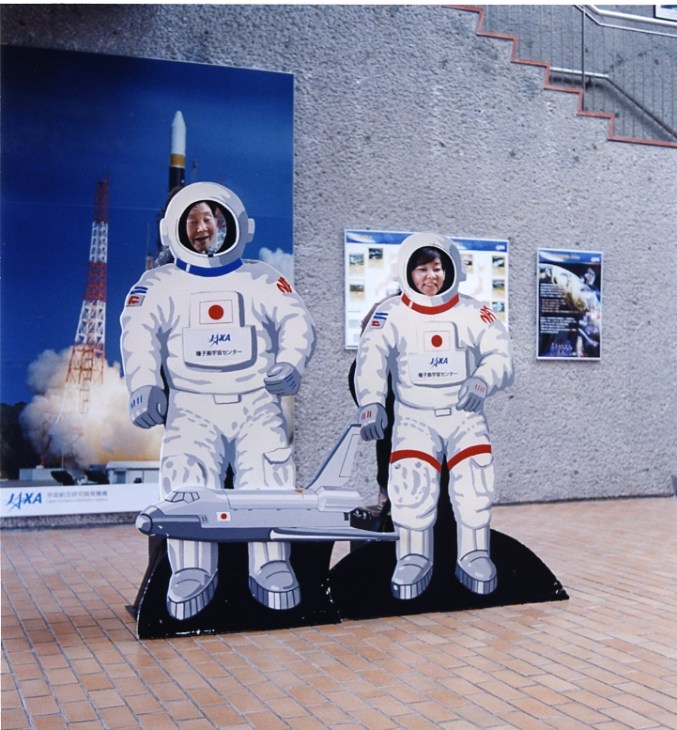 Two people pose with life-size cardboard cutouts of Japanese astronauts in white spacesuits at the space museum.