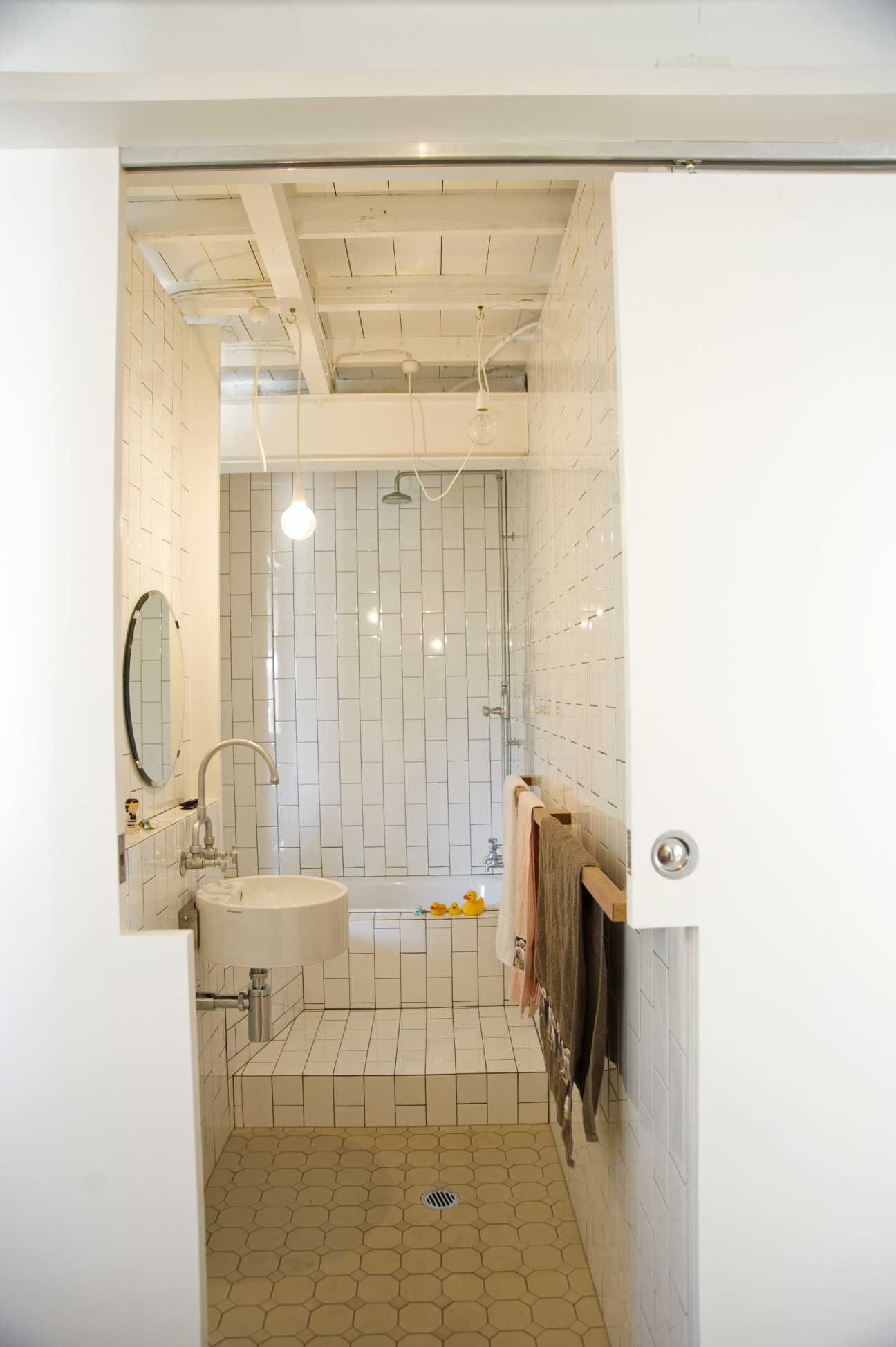 Bathroom with white subway tile walls, exposed wooden ceiling beams, round sink, circular mirror, and butchers' tiles on the floor.