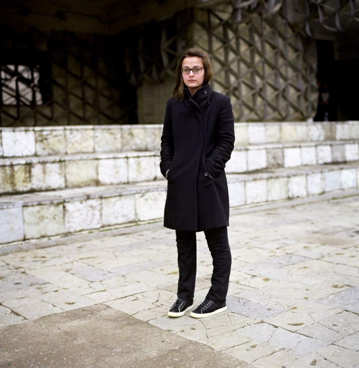 Young woman in black coat and sneakers standing in Pristina.