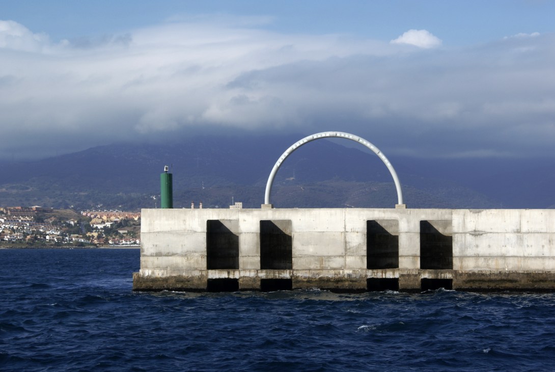 White illuminated arch beacon marking Barcelona's harbor entrance with coastal city and mountains in background.