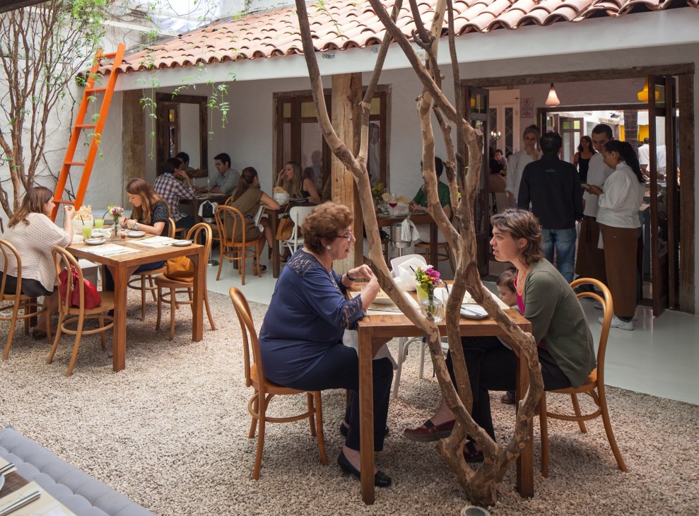 Restaurant courtyard with diners seated at wooden tables under gnarled tree branches, white walls and terracotta roof.