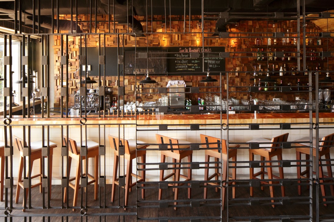 Modern restaurant interior with wooden chairs, brick wall backdrop, and a metal-framed counter with glass panels.