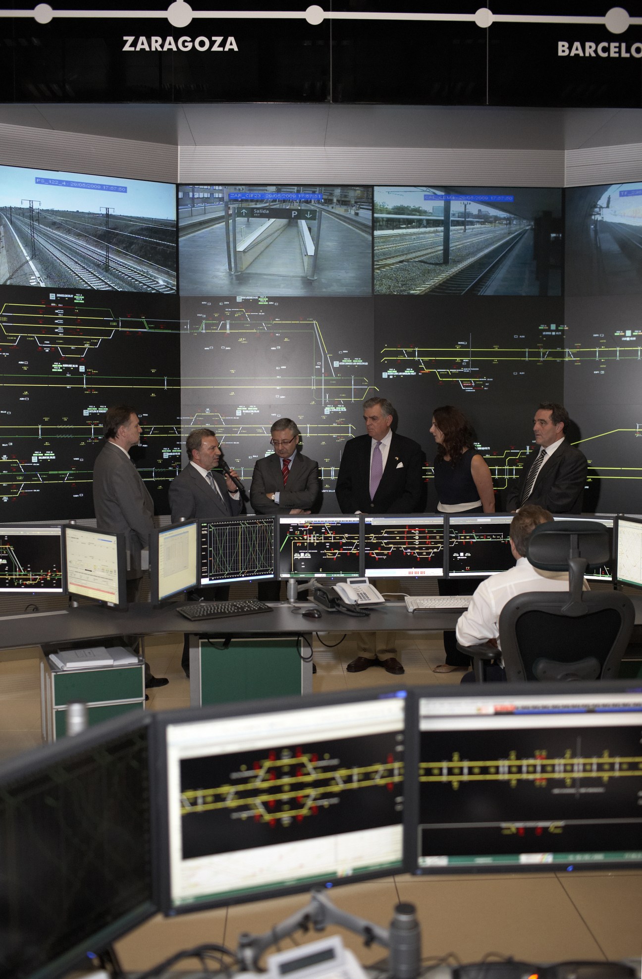 Officials briefing in a rail command center with monitors displaying train routes and surveillance feeds.