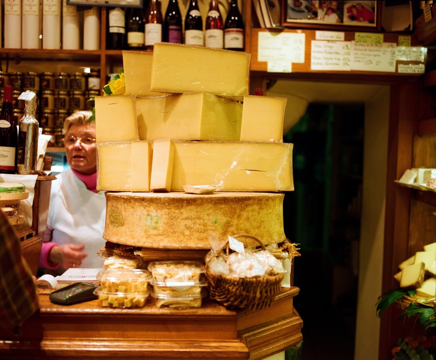 Stacked wheels of yellow cheese displayed in a Paris fromagerie with wine bottles and shopkeeper in background.