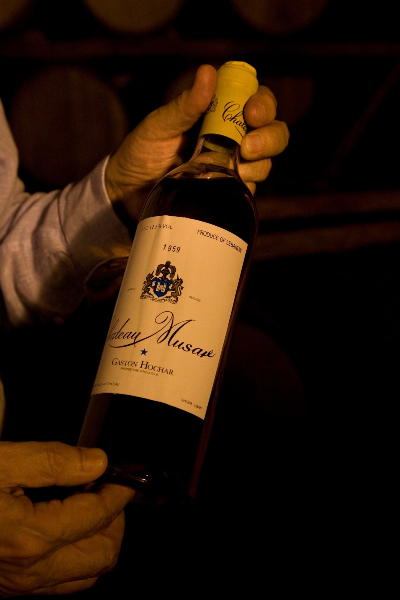 A hand holds a bottle of Château Musar 1959 wine with a yellow foil cap in dim lighting.