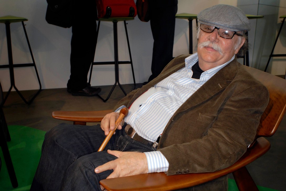 Brazilian architect Sergio Rodrigues sits on a wooden chair holding an axe handle, surrounded by modern furniture at ClassiCon.