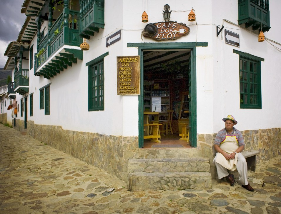 Owner Eloy sits outside Cafe don Eloy, a colonial-style coffee shop with white walls and green wooden doors and shutters on a cobblestone street in Villa de Leyva.
