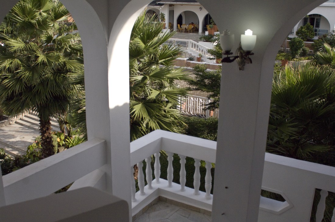 White colonial-style arches and balustrade with palm trees and courtyard visible through openings at Cap Kivu Hotel.
