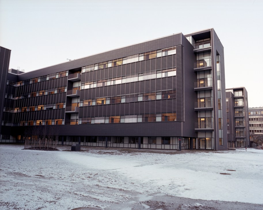Modern hospital building with dark metal cladding and rhythmic rows of windows, photographed across a snowy courtyard.