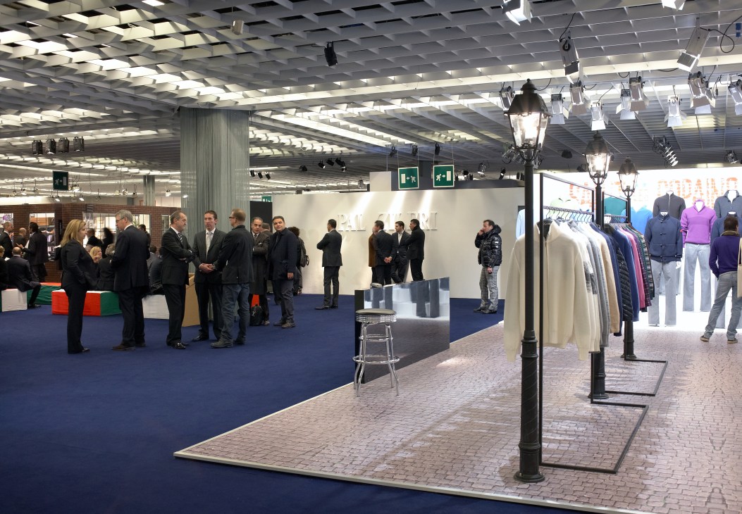 Interior of Pitti Immagine Uomo exhibition hall with visitors browsing clothing displays and purple garments on mannequins.