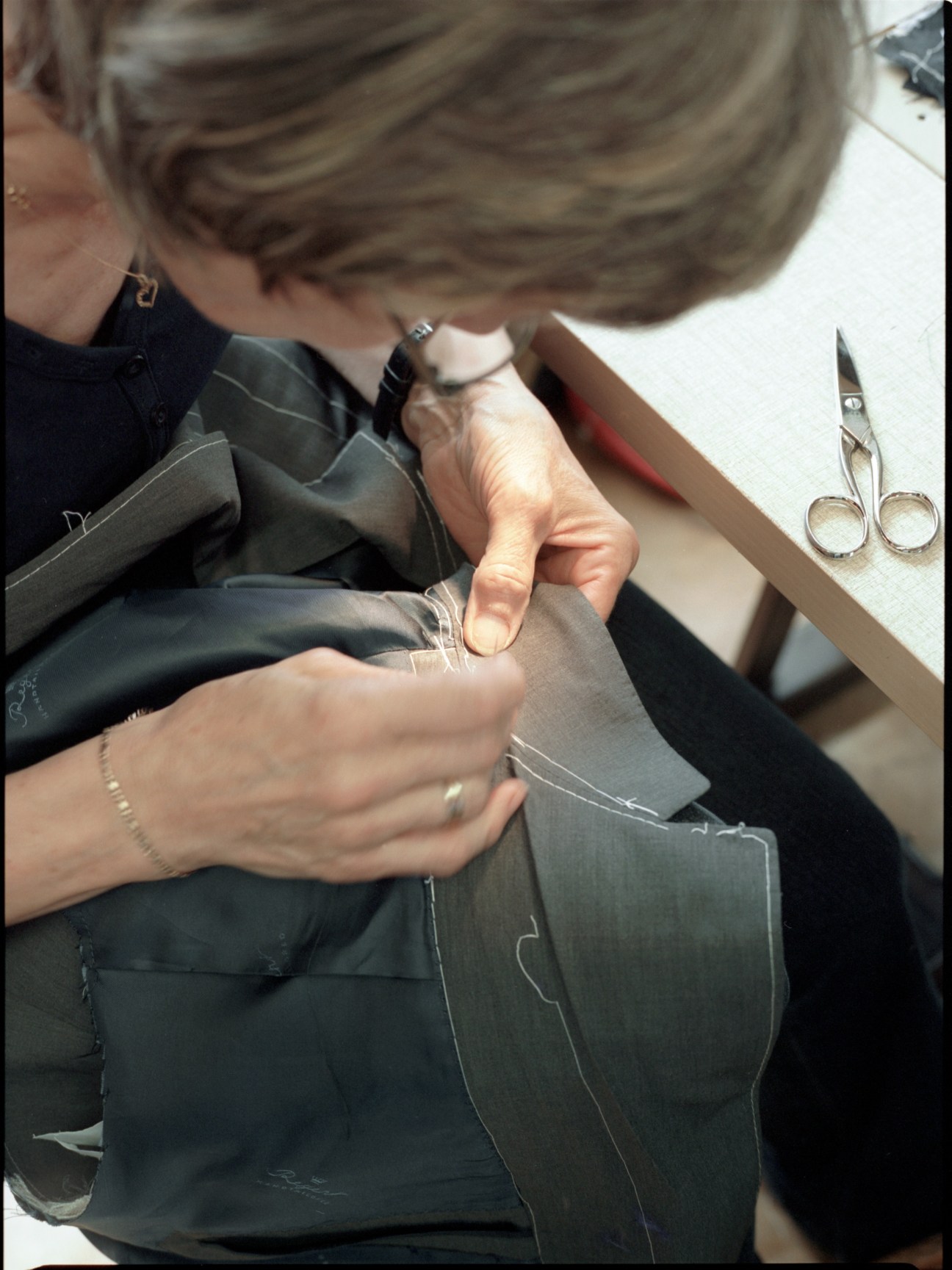 Tailor's hands sewing complicated seams by hand on dark fabric with scissors nearby.