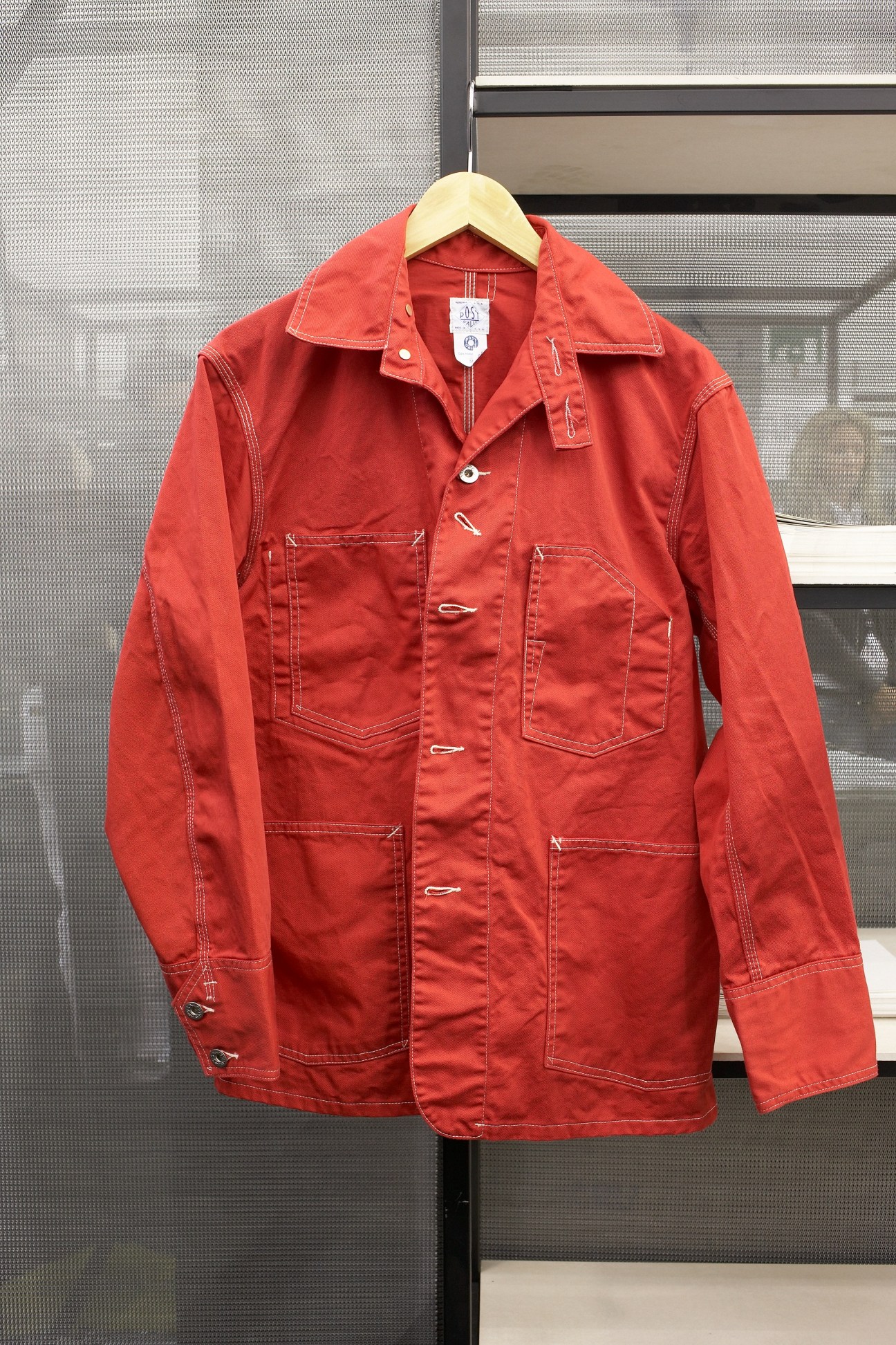 Red cotton engineer's jacket by Post O'Alls on wooden hanger against mesh background.