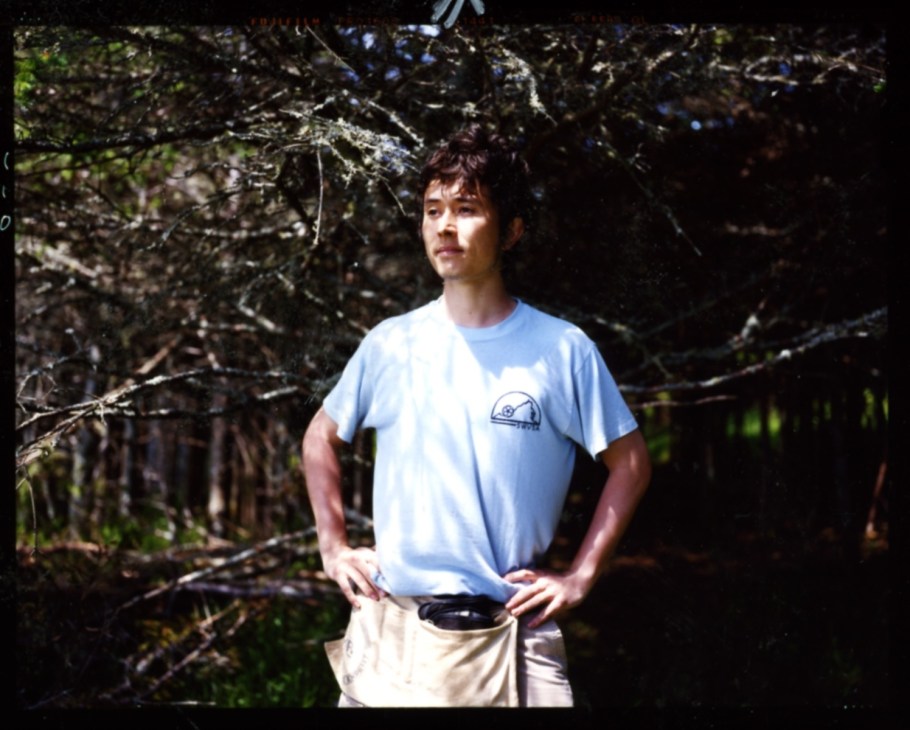 Architect Philippe Mizutani wearing a light blue t-shirt stands among trees during Ghost Laboratory camp.