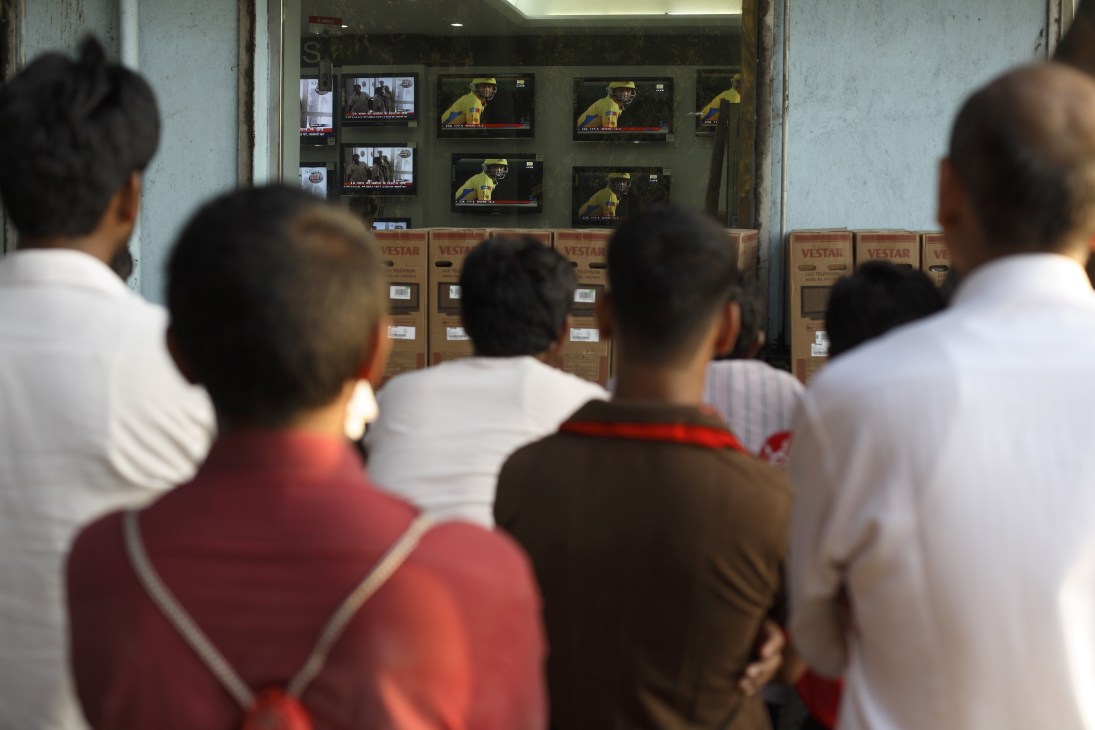 People gathered outside a TV shop watching multiple screens displaying cricket match footage.
