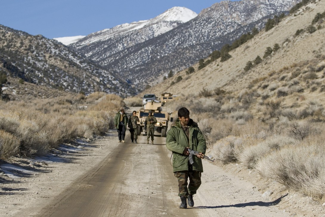 Soldier sweeps desert road for explosives with military convoy and snow-capped mountains in background.