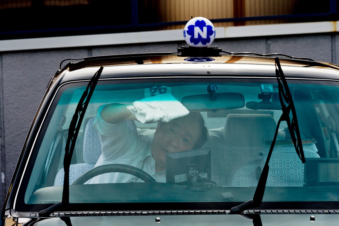 A Nihon Kotsu taxi driver in white gloves cleaning the interior of a taxi with embroidered seat covers.