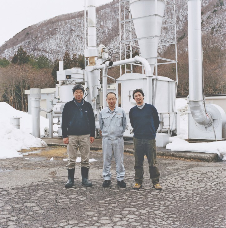 Three men stand in front of industrial equipment with snowy mountains in the background.