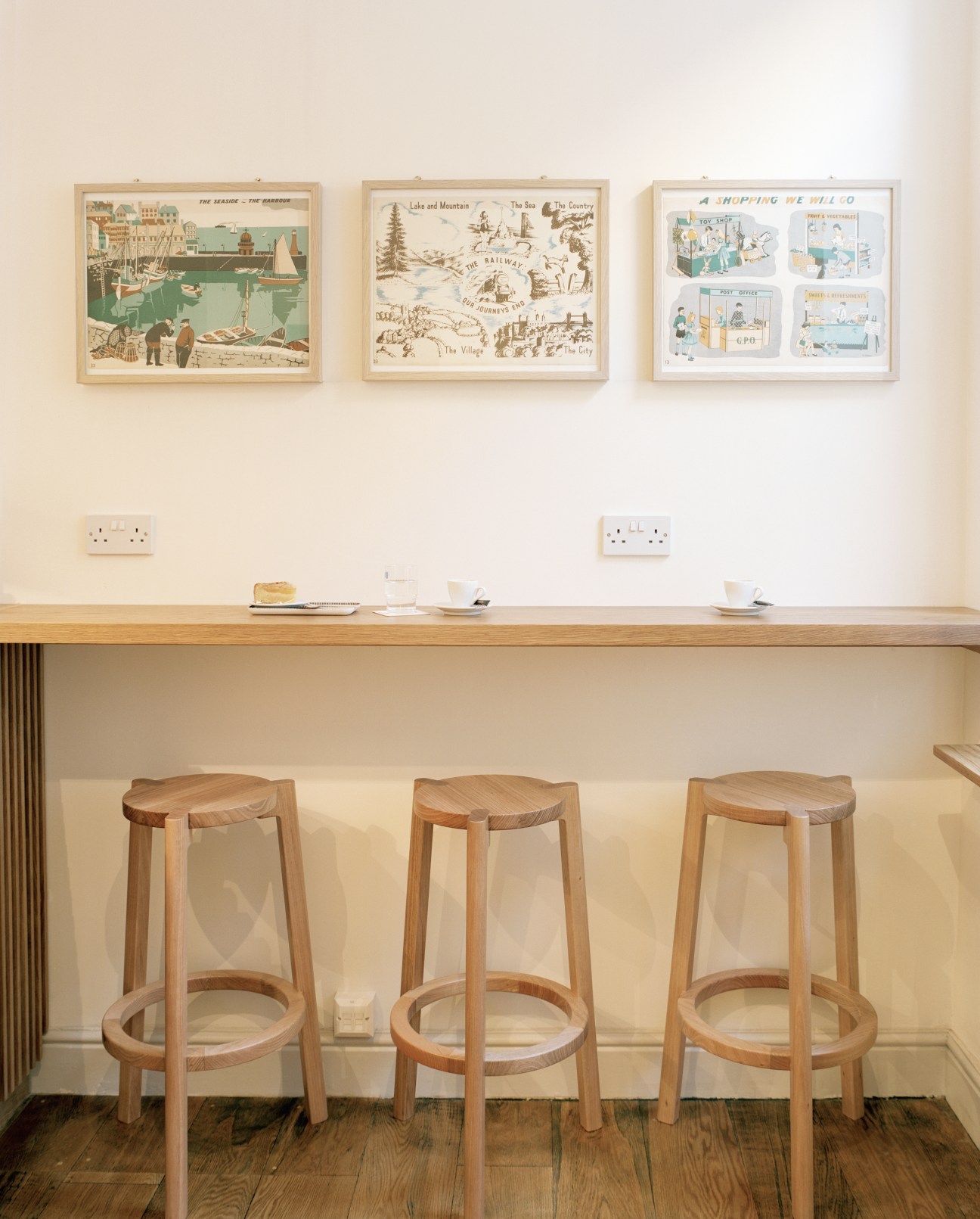 Three wooden bar stools by One/Third positioned at a counter beneath framed vintage maps on a cream wall.