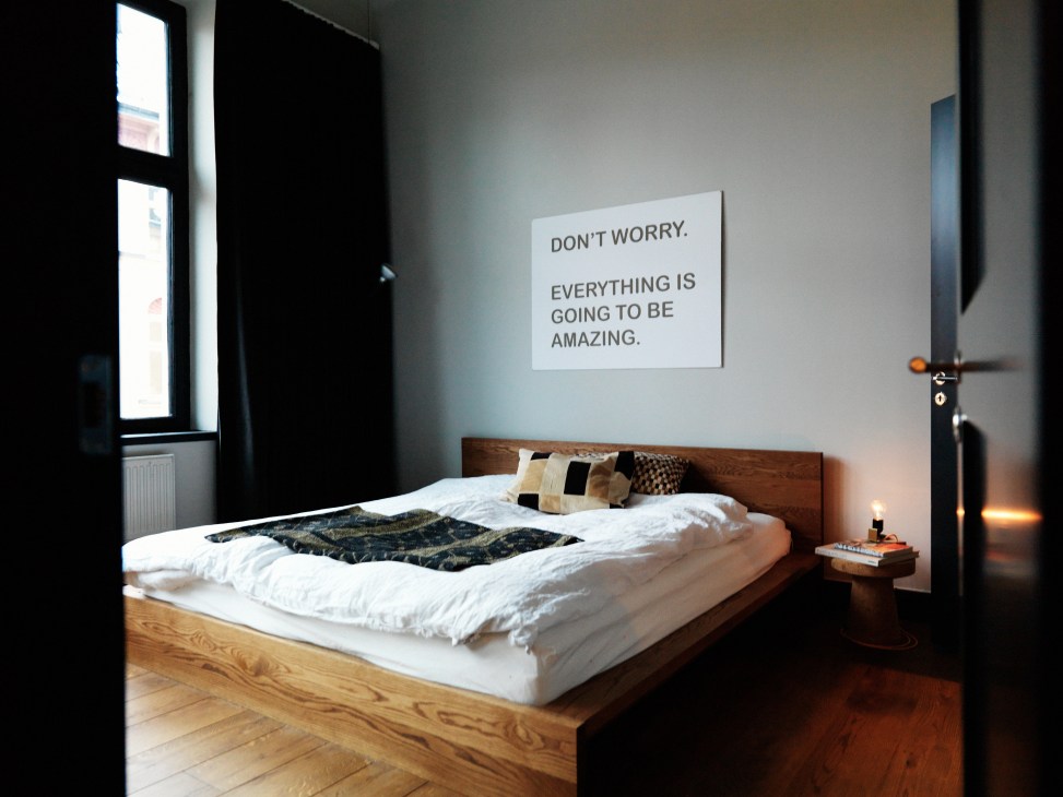 Minimalist bedroom with wooden bed frame, white bedding, and motivational wall text.