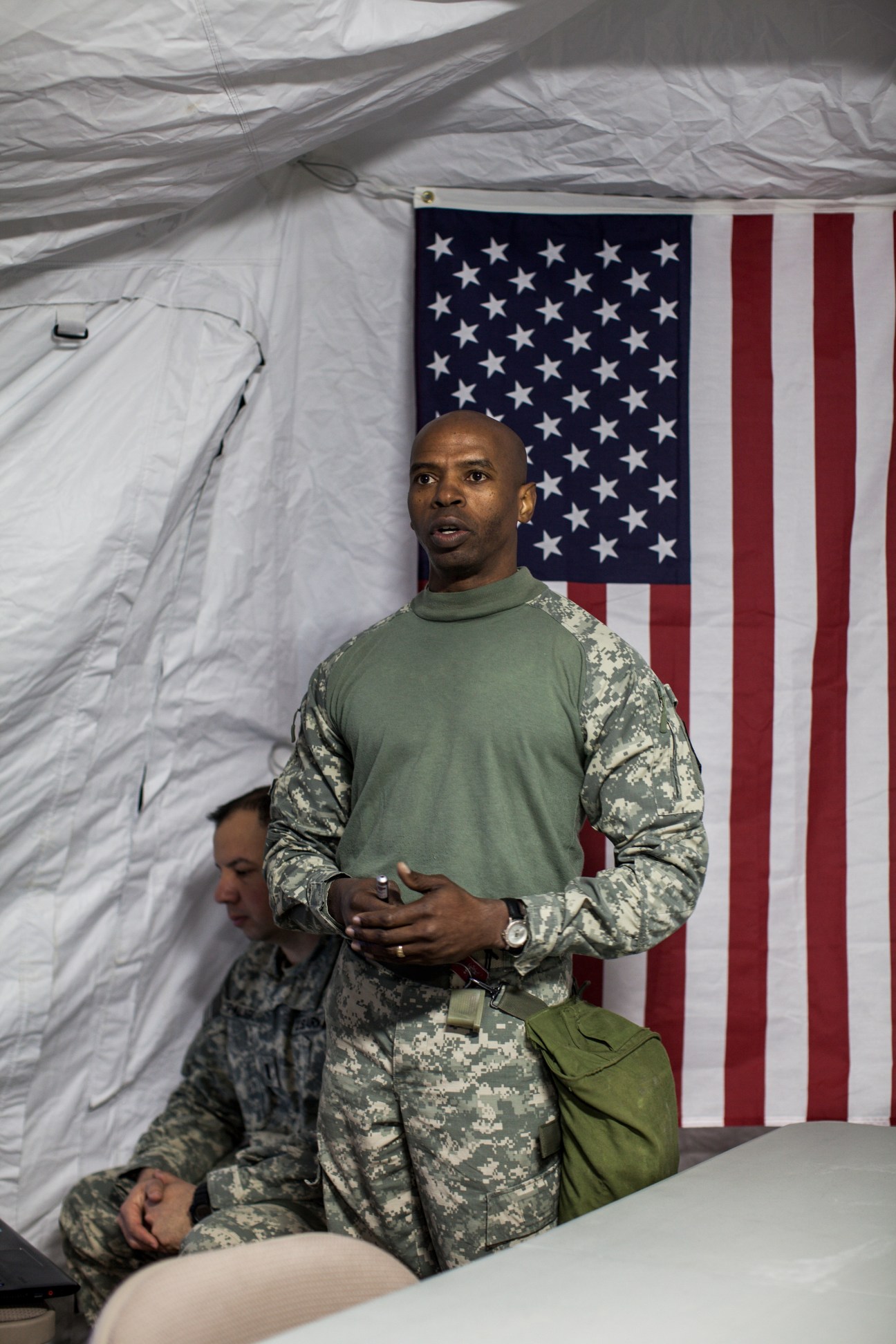 US military officer in camouflage uniform standing in front of American flag inside tent