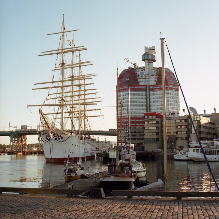 Tall ship with white hull and golden masts docked in Gothenburg harbour beside modern waterfront buildings.