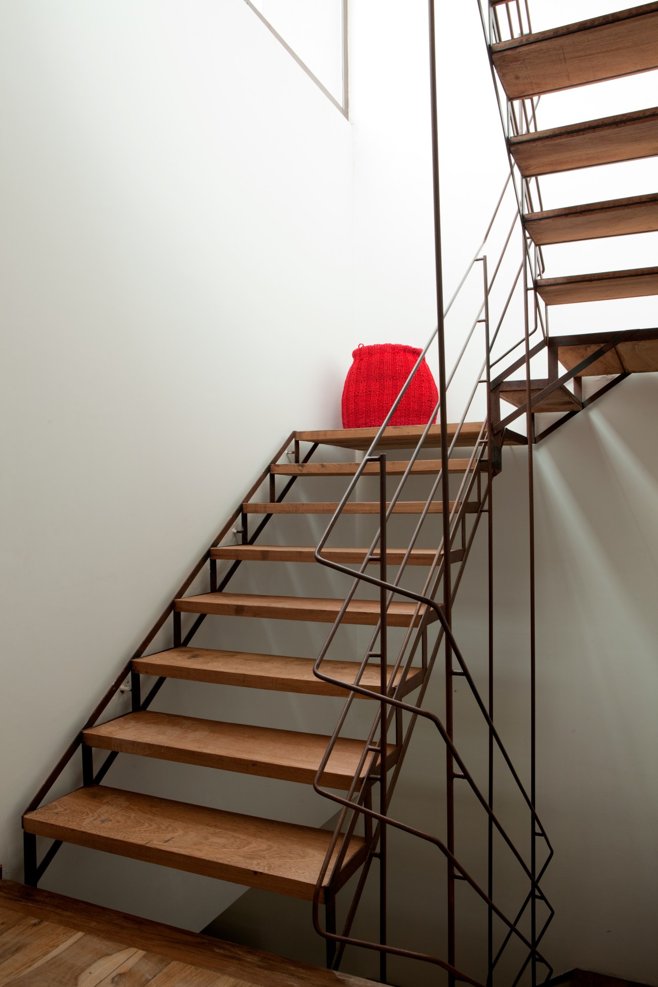 Industrial-style wooden staircase with metal railings and a red cushioned chair against white walls beneath natural skylight.