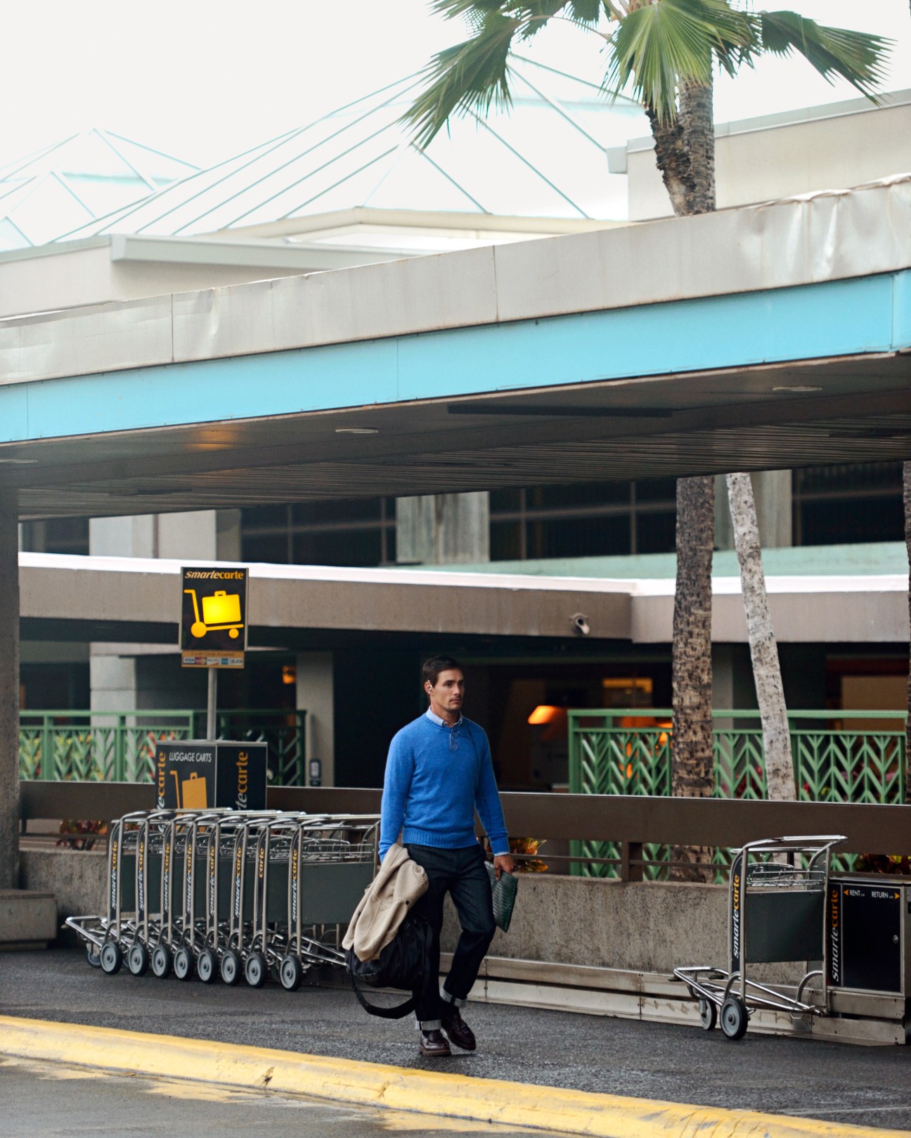 Man in blue jumper and dark trousers carries coat outside modern airport terminal with palm tree.