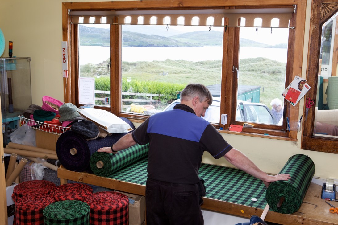 Don Mackay works at a loom in his home with Harris Tweed cloth rolls visible, overlooking a beach landscape through large windows.