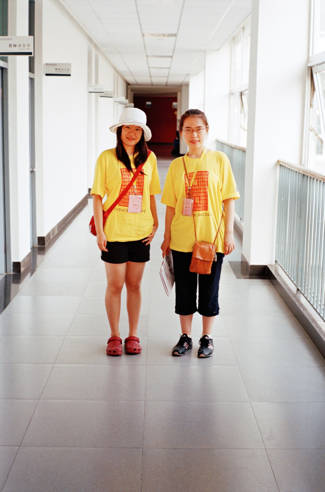 Two young women in yellow Chinese Bridge camp t-shirts and lanyards stand in a bright hallway.