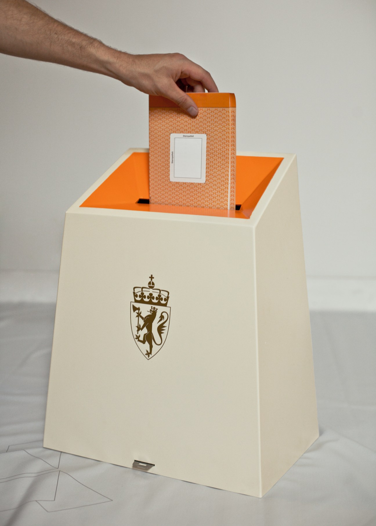Hand inserting orange patterned ballot paper into a cream-colored Norwegian ballot box with golden lion emblem.