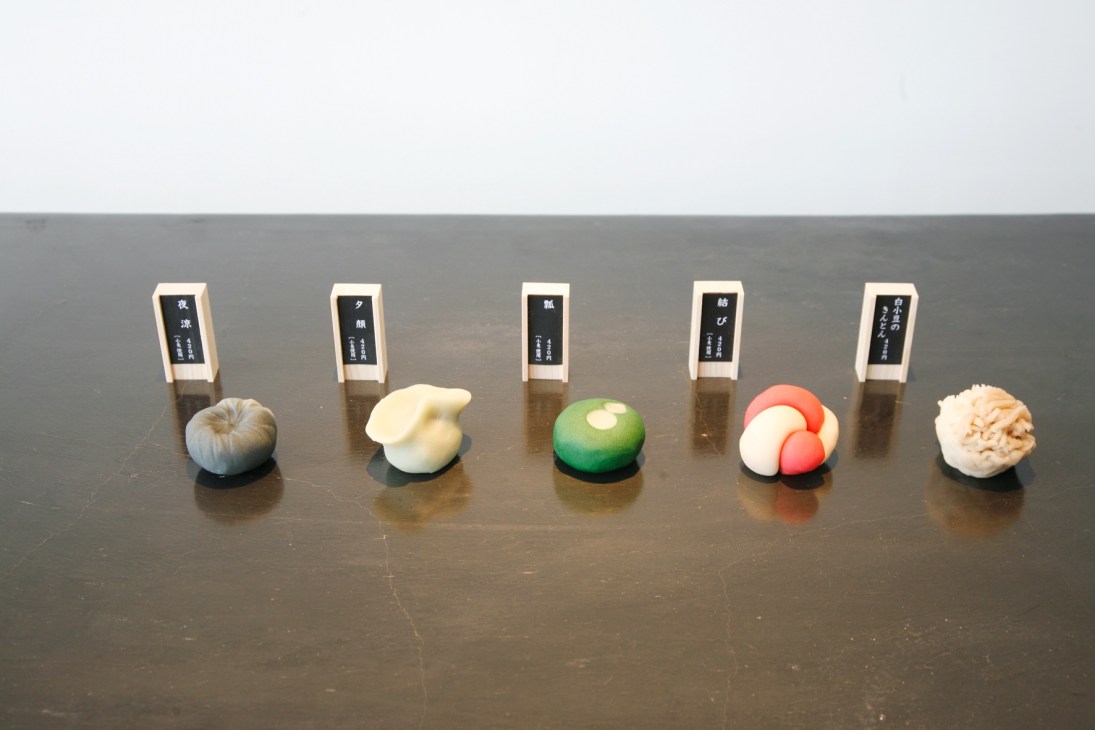 Five colorful Japanese sweets displayed with labeled wooden cards on a dark surface at Baishinka, Tokyo.