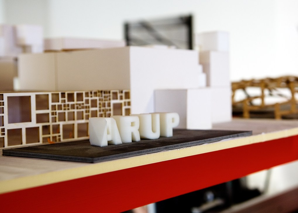 Architectural models displayed on a red table, including white building blocks and a latticed model structure.