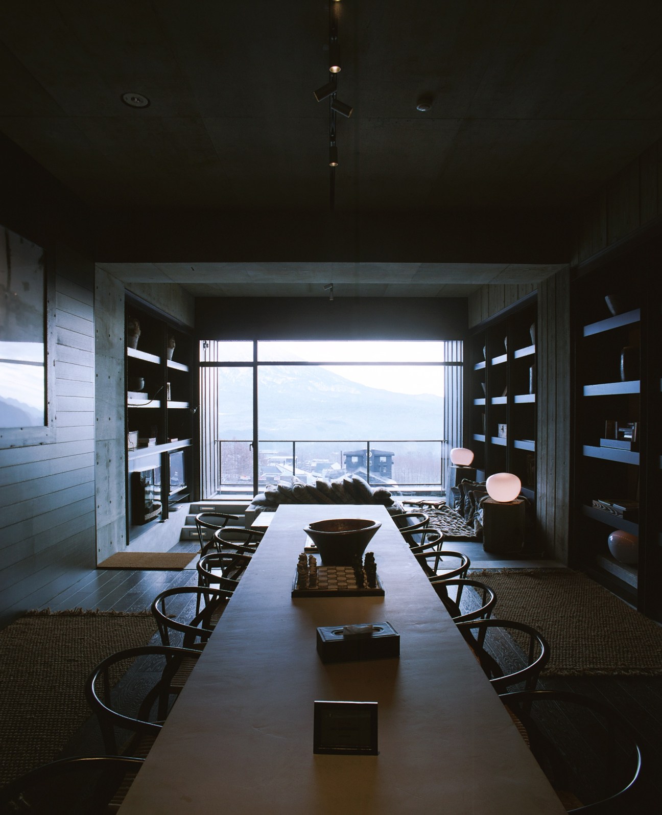 Modern interior of Suiboku residence with long table, chairs, built-in shelving and large windows overlooking snowy mountains.
